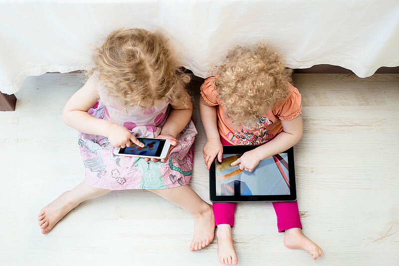 Zwei kleine Kinder mit lockigem Haar sitzen auf dem Boden und benutzen jeweils ein digitales Gerät. Ein Kind hält ein Smartphone in der Hand, während das andere ein Tablet benutzt. Beide sind auf ihre Bildschirme konzentriert und sitzen nebeneinander neben einem Bett.